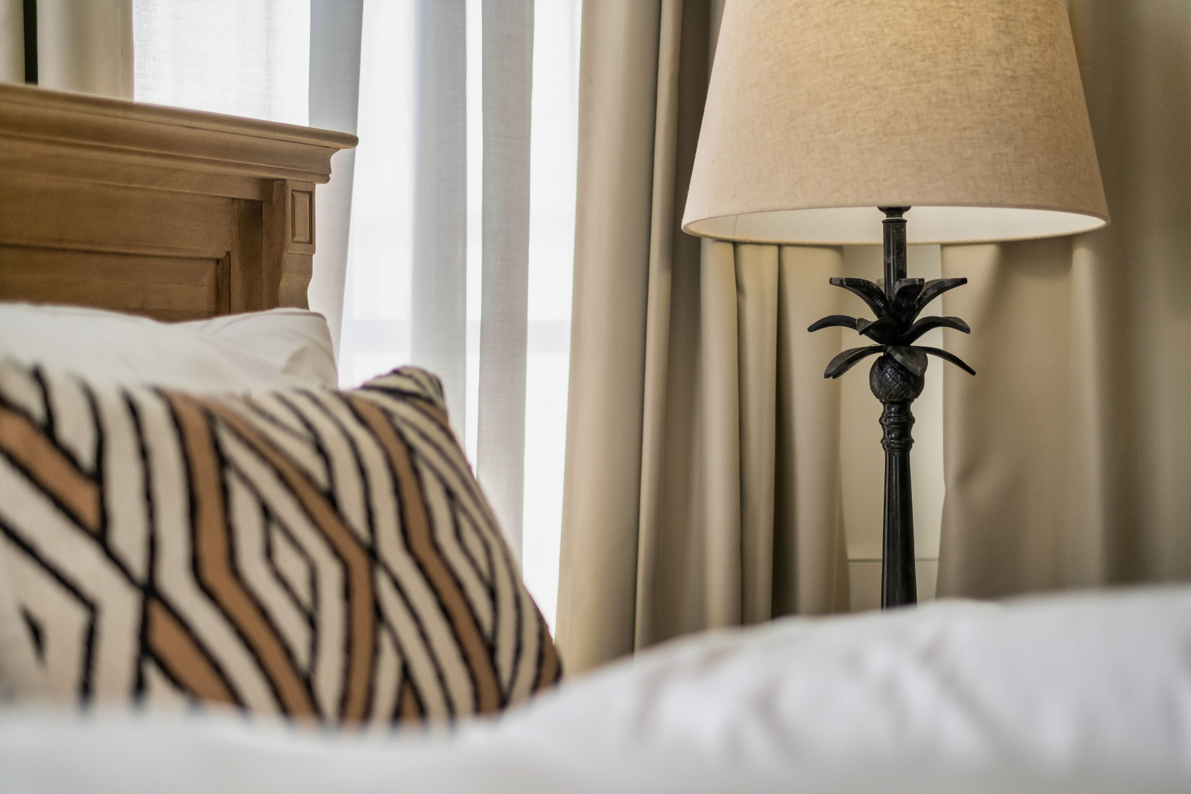 A bed with a patterned pillow, a lamp with a beige lampshade, and curtains in a hotel room.