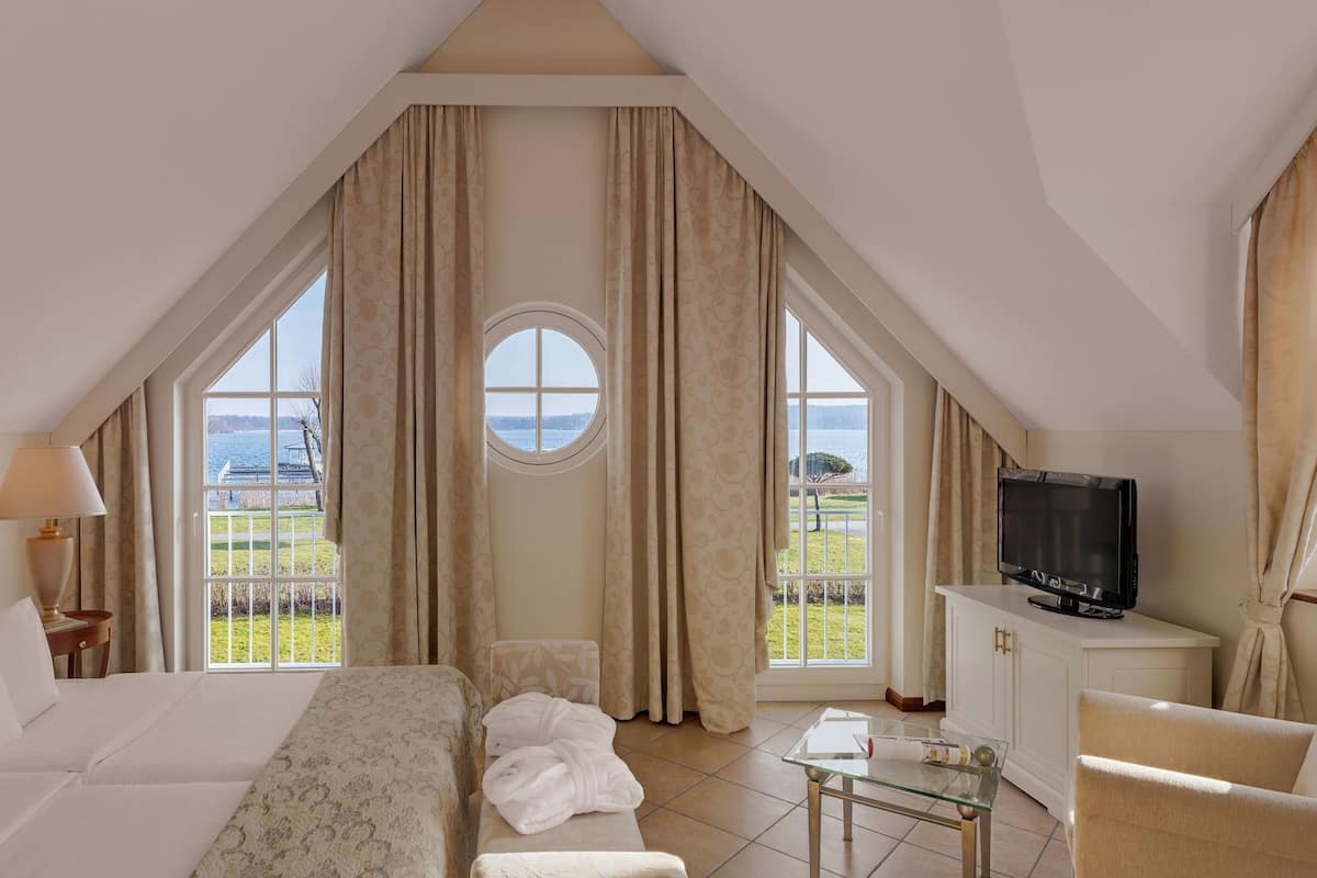 A bedroom with a large bed, a TV, a glass table, and a view of a lake through a window.