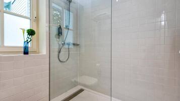 A glass-enclosed shower with a handheld showerhead, a white tiled wall, and a window with a plant.