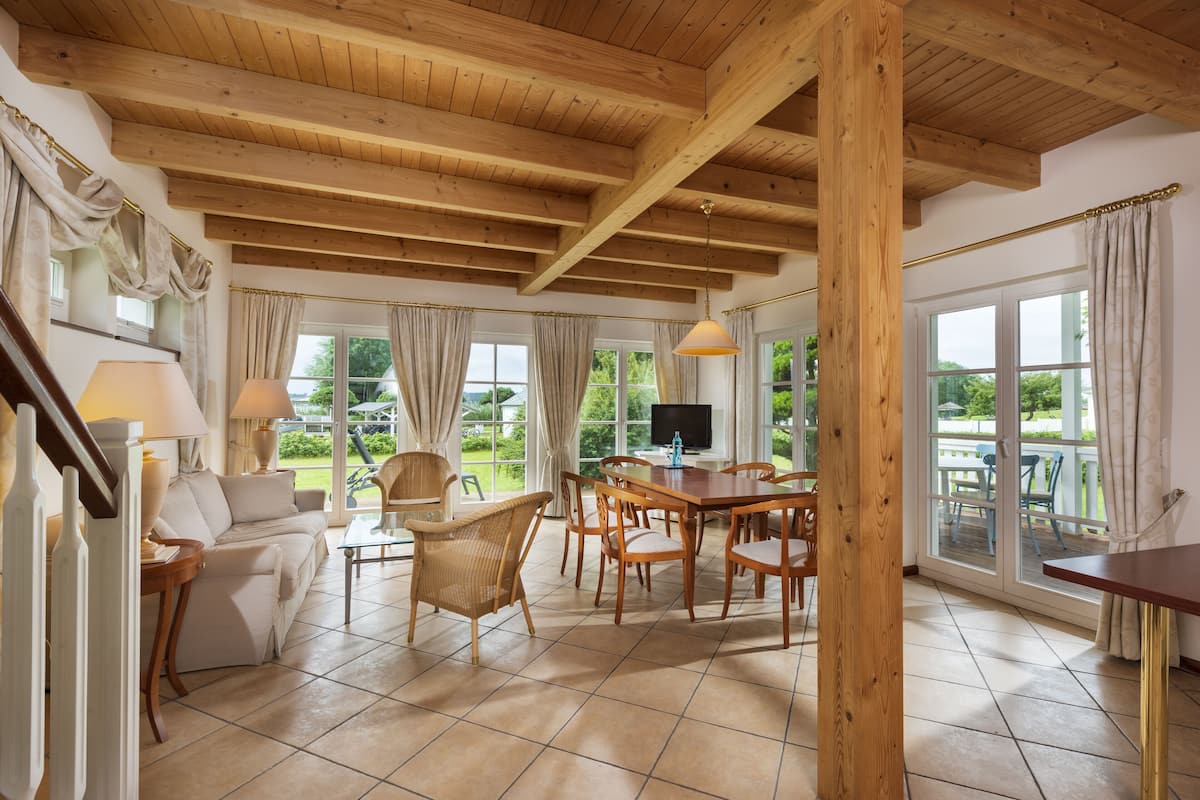 A spacious living room with a wooden ceiling, large windows, a dining area with wooden furniture, and a view of the outdoors.