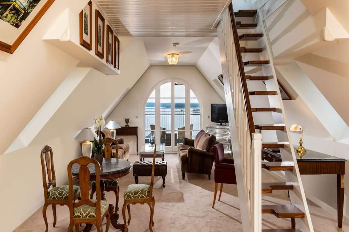 A spacious loft with a high ceiling, a dining area with a round table and chairs, a cozy seating area with sofas, a staircase, and a large arched window offering a view.