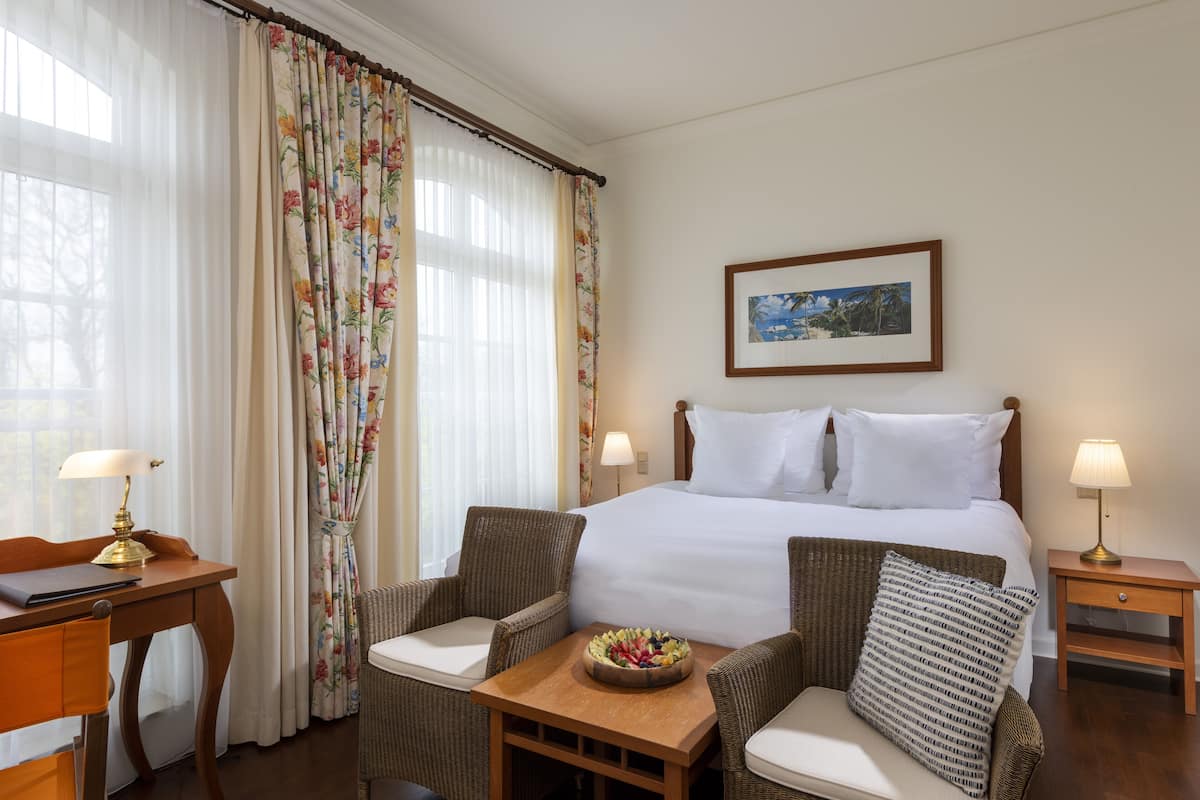 A hotel room with a bed, two chairs, a small table with a fruit bowl, a nightstand with a lamp, and a framed picture on the wall.