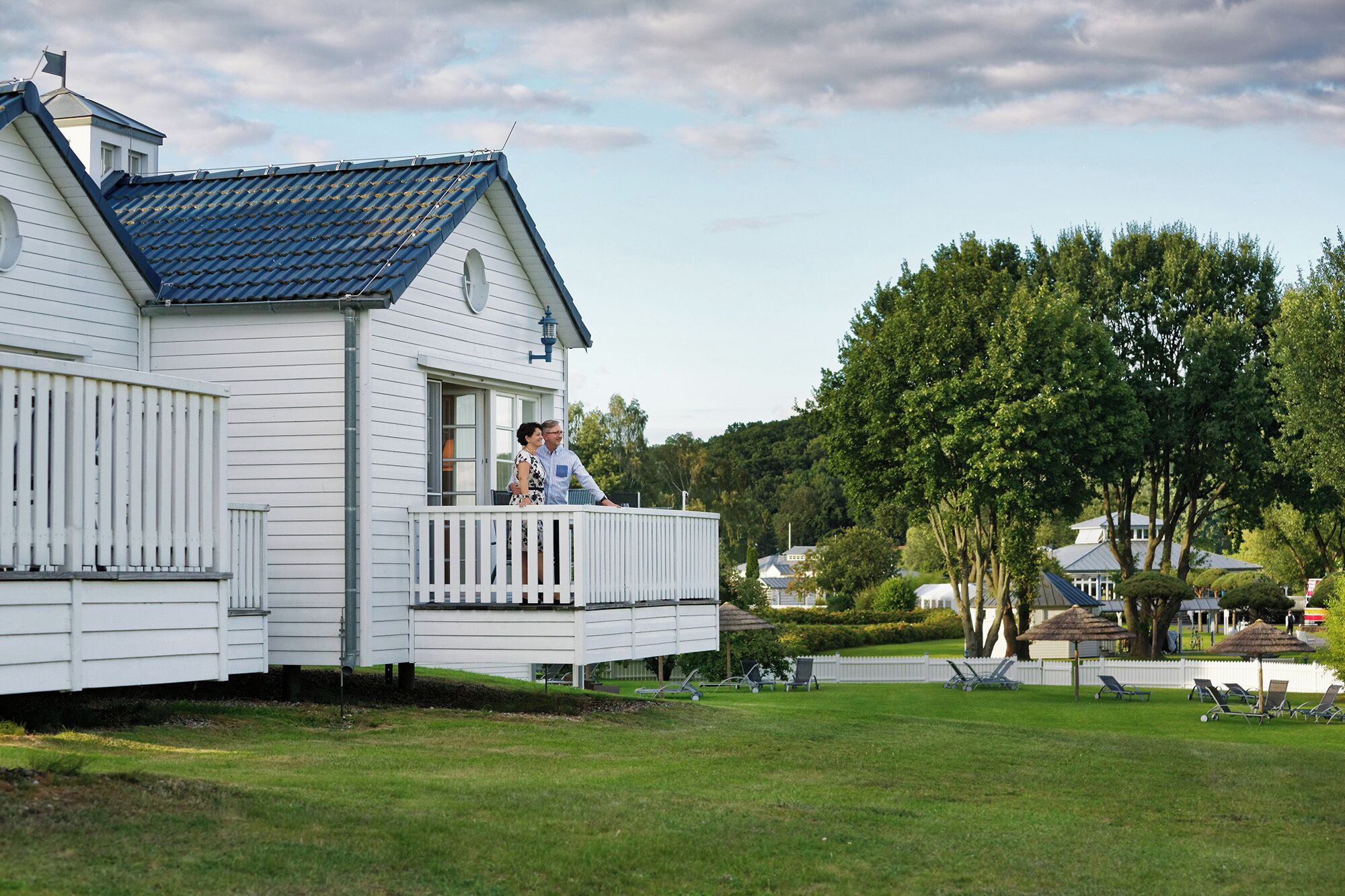 A white house with a blue roof, a balcony, and a well-maintained lawn with outdoor furniture.