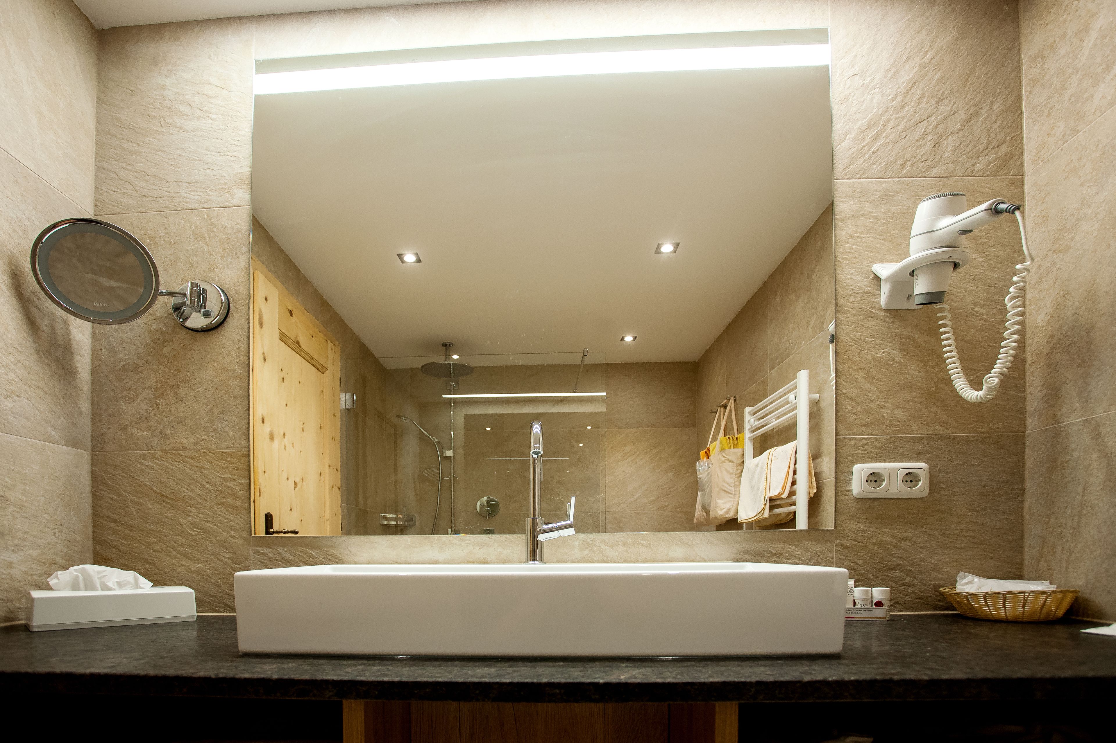 Un baño moderno con un espejo grande, un lavamanos, un cabezal de ducha de mano y un perchero para toallas.
