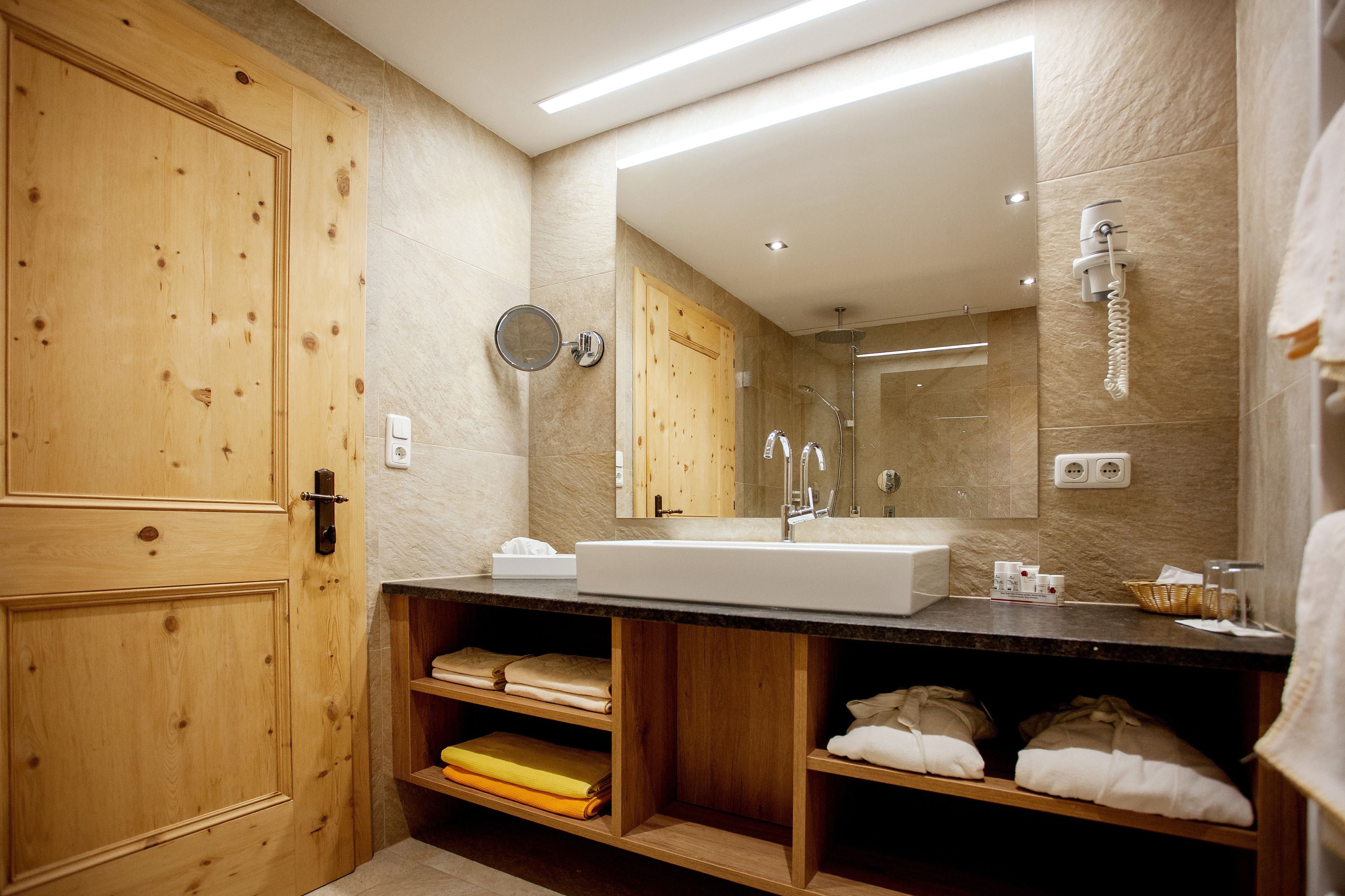 A modern bathroom with a wooden vanity, a large mirror, a shower, and built-in shelves for towels.