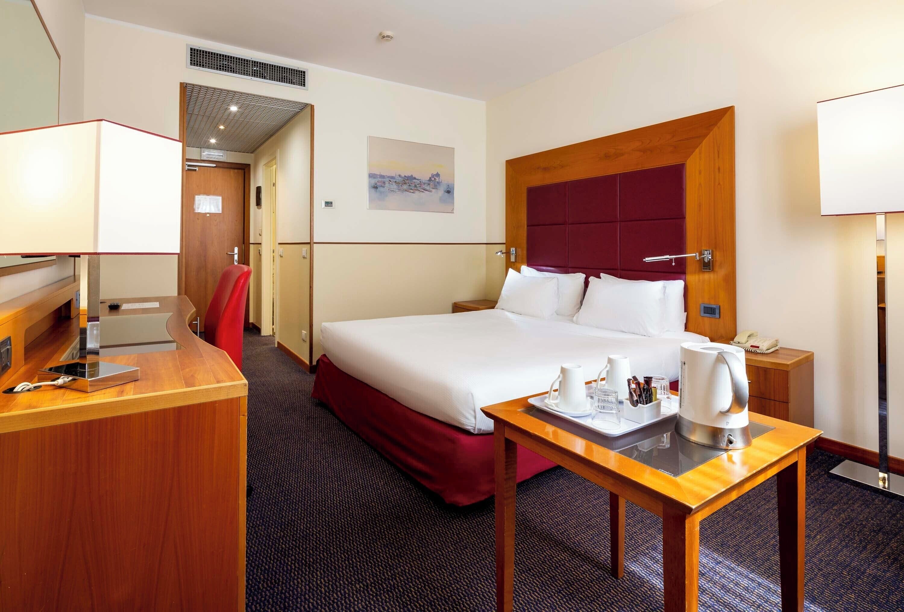 A hotel room with a bed, a desk with a telephone, a bedside table with a kettle, and a painting on the wall.