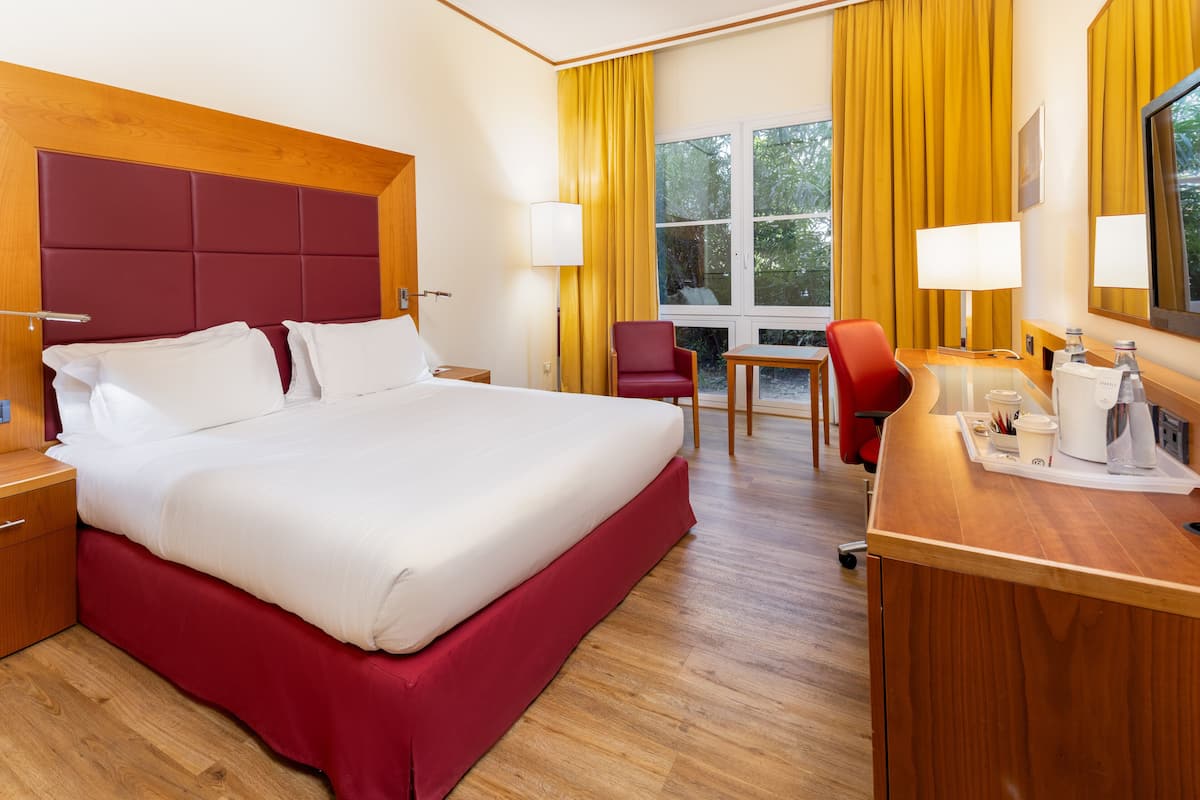 Ein Hotelzimmer mit einem großen Bett, einem Schreibtisch mit Stuhl, einem Fenster mit Vorhängen und einem hölzernen Kopfteil.