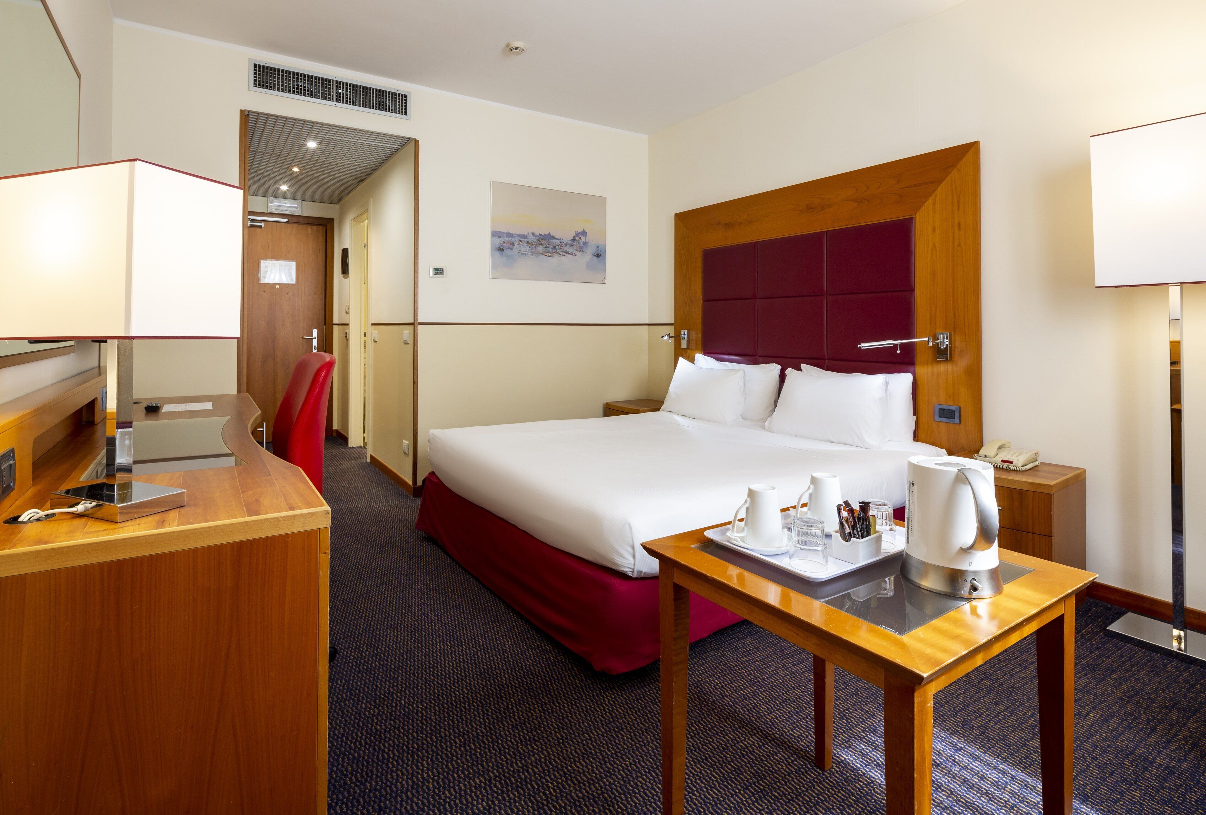 A hotel room with a large bed, a desk with a lamp, a red chair, a bedside table with a phone and a kettle, and a painting on the wall.