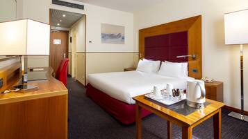 A hotel room with a large bed, a desk with a lamp, a red chair, a bedside table with a phone and a kettle, and a painting on the wall.