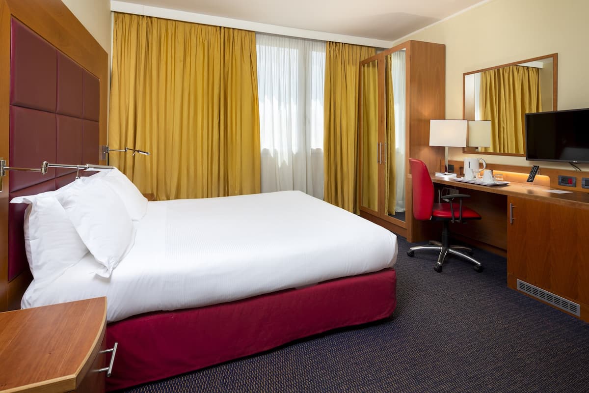 Ein Hotelzimmer mit einem großen Bett, einem Schreibtisch mit Stuhl, einem Fernseher und einem Fenster mit Vorhängen.
