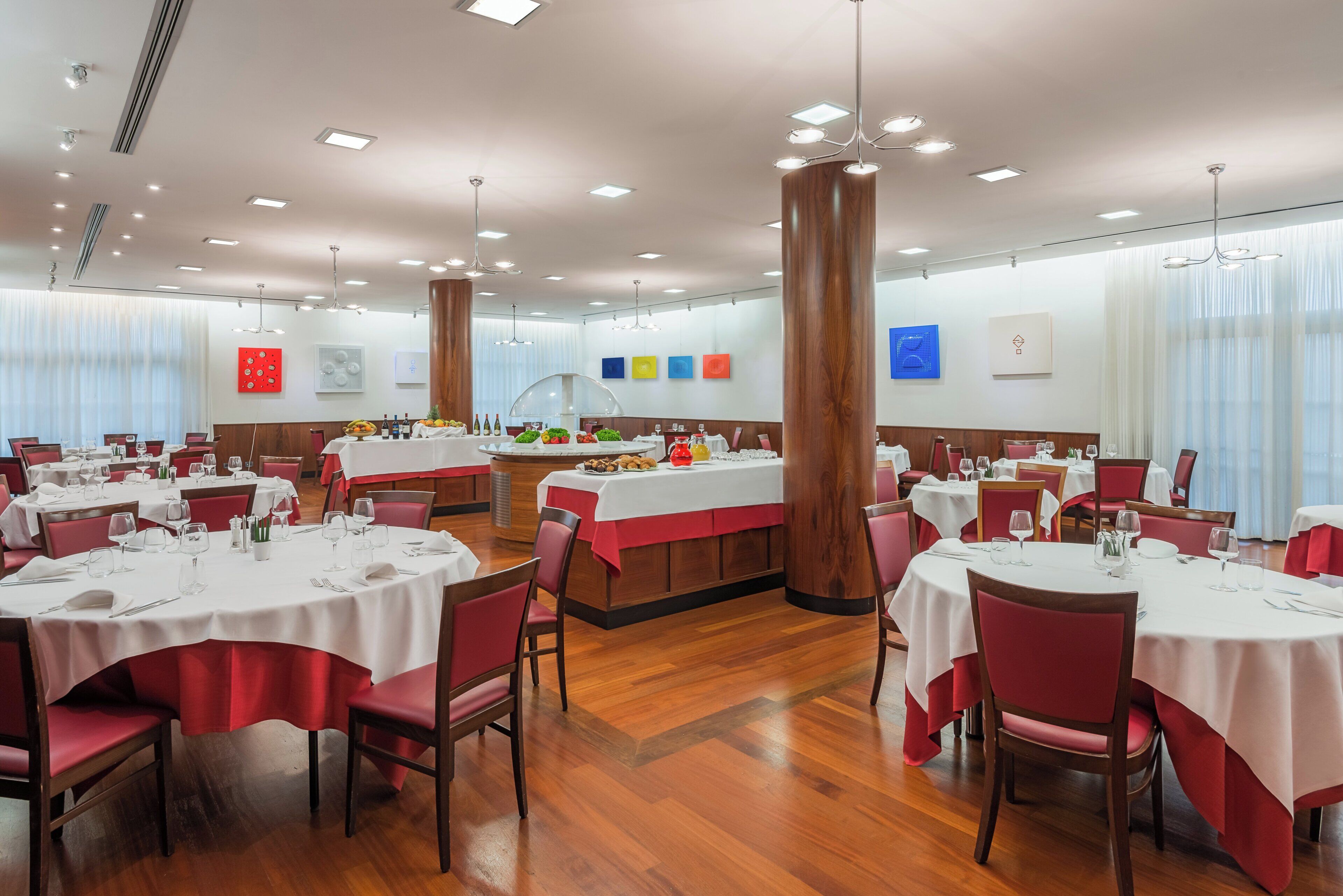 A spacious dining area with multiple round tables, red chairs, and a central island with food and drinks.