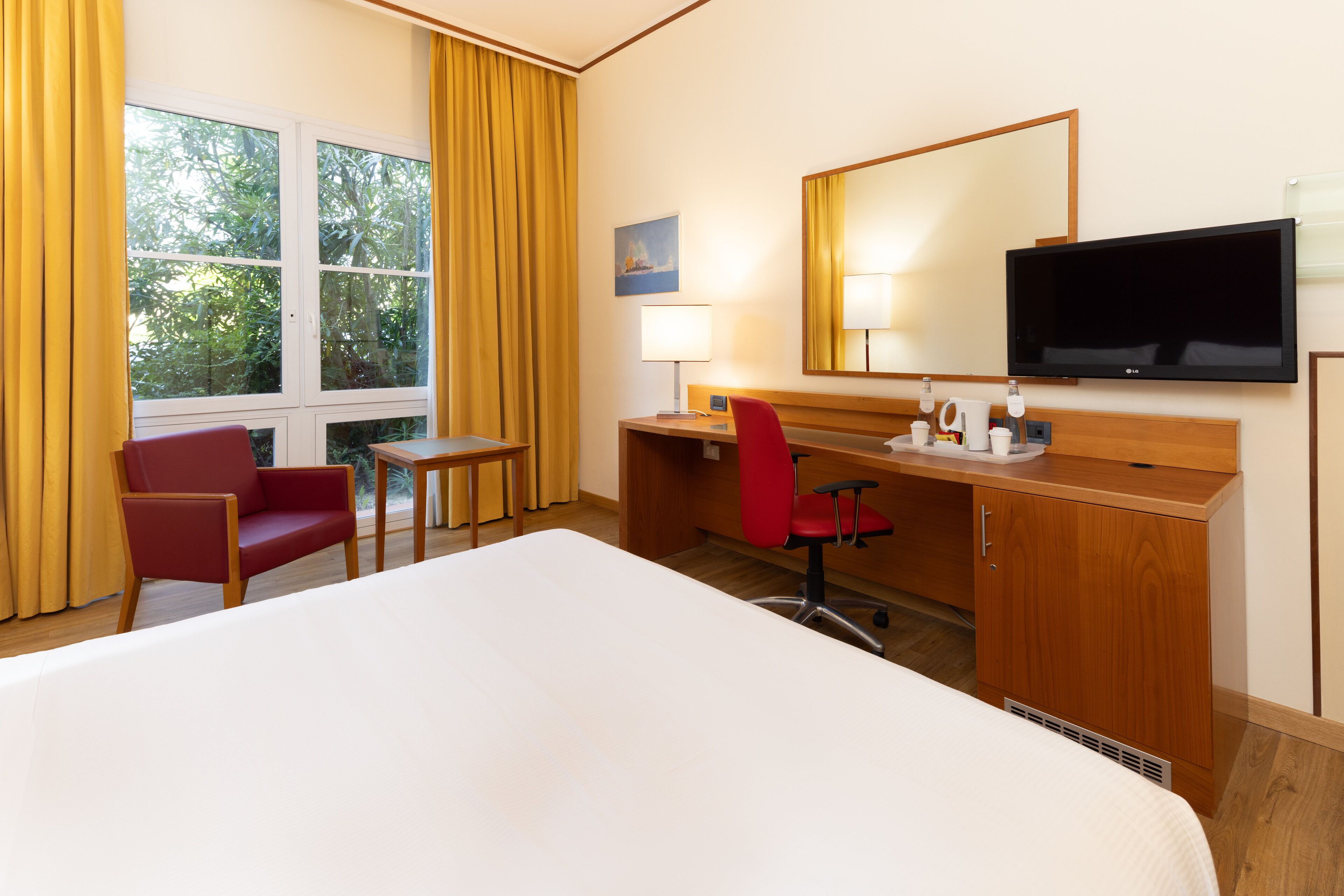 Quarto de hotel com cama, escrivaninha, cadeira, televisão e janela com cortinas.