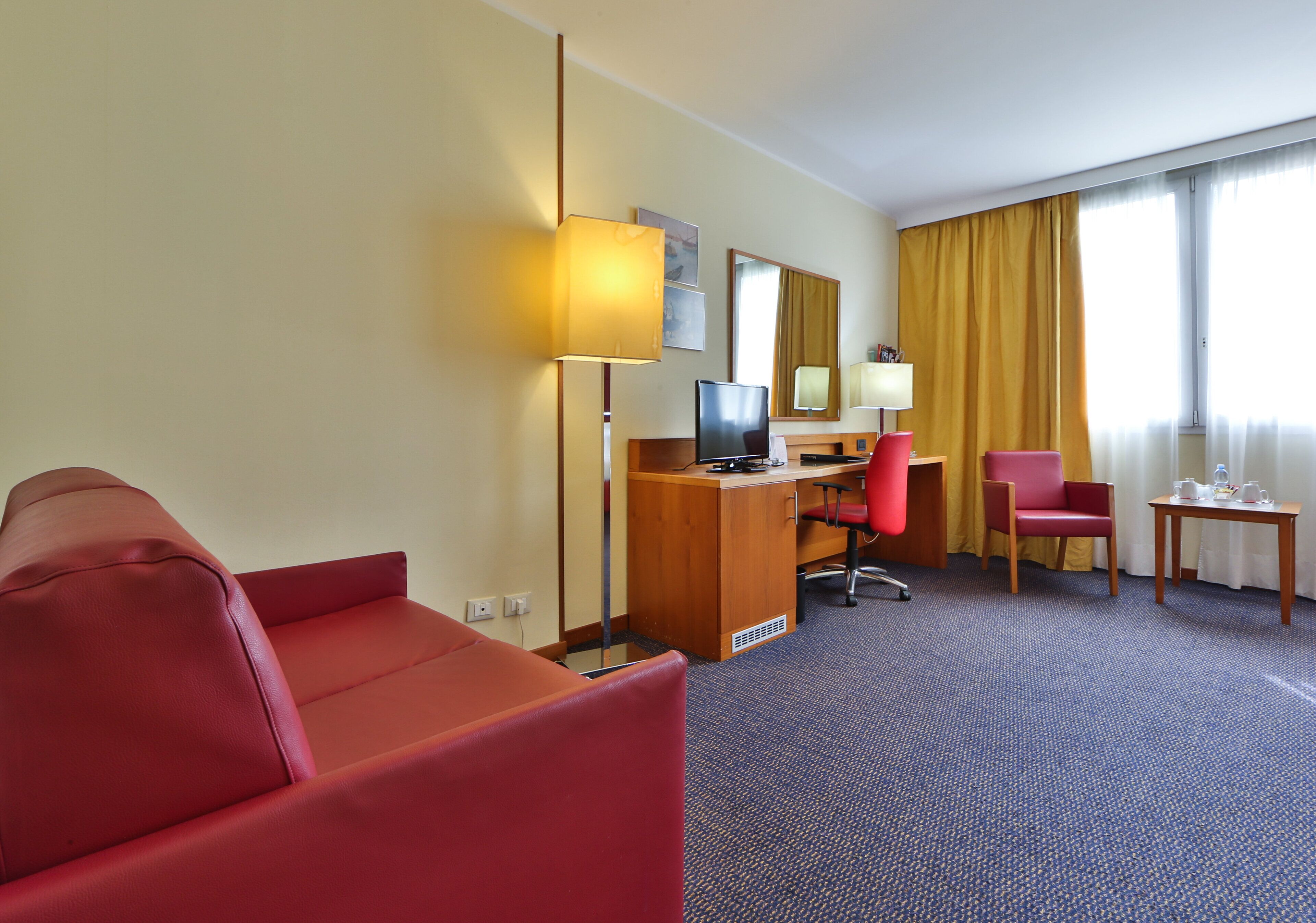 Habitación de hotel con sofá rojo, un escritorio de madera, una computadora, una silla de oficina roja y una pequeña mesa con una lámpara.