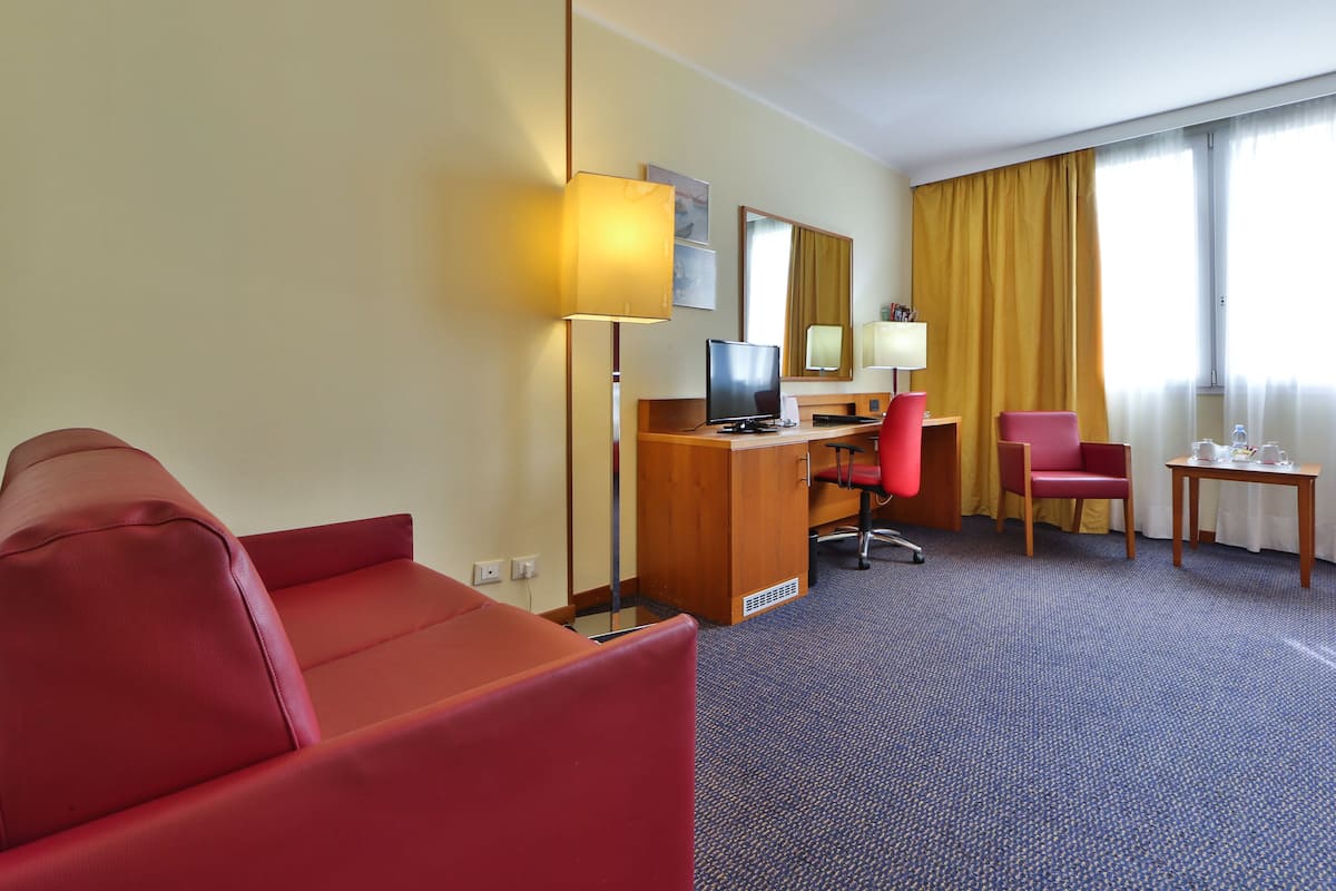 Une chambre d’hôtel avec un canapé rouge, un bureau en bois, un ordinateur, un fauteuil de bureau rouge et une petite table avec une lampe.