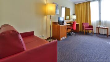 Habitación de hotel con sofá rojo, un escritorio de madera, una computadora, una silla de oficina roja y una pequeña mesa con una lámpara.