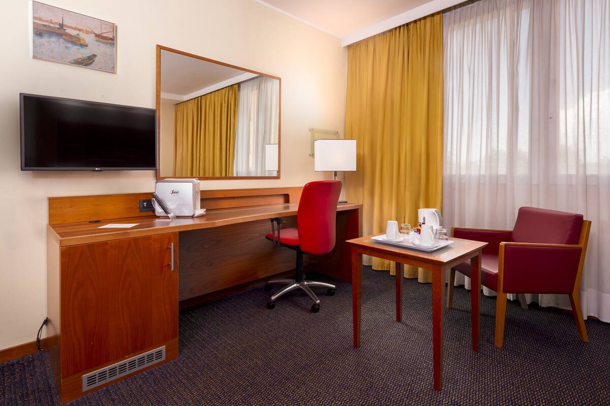 Ein Hotelzimmer mit einem Holztisch, einem roten Bürostuhl, einem Fernseher, einem Spiegel, einem Fenster mit gelben Vorhängen und einem roten Sessel.