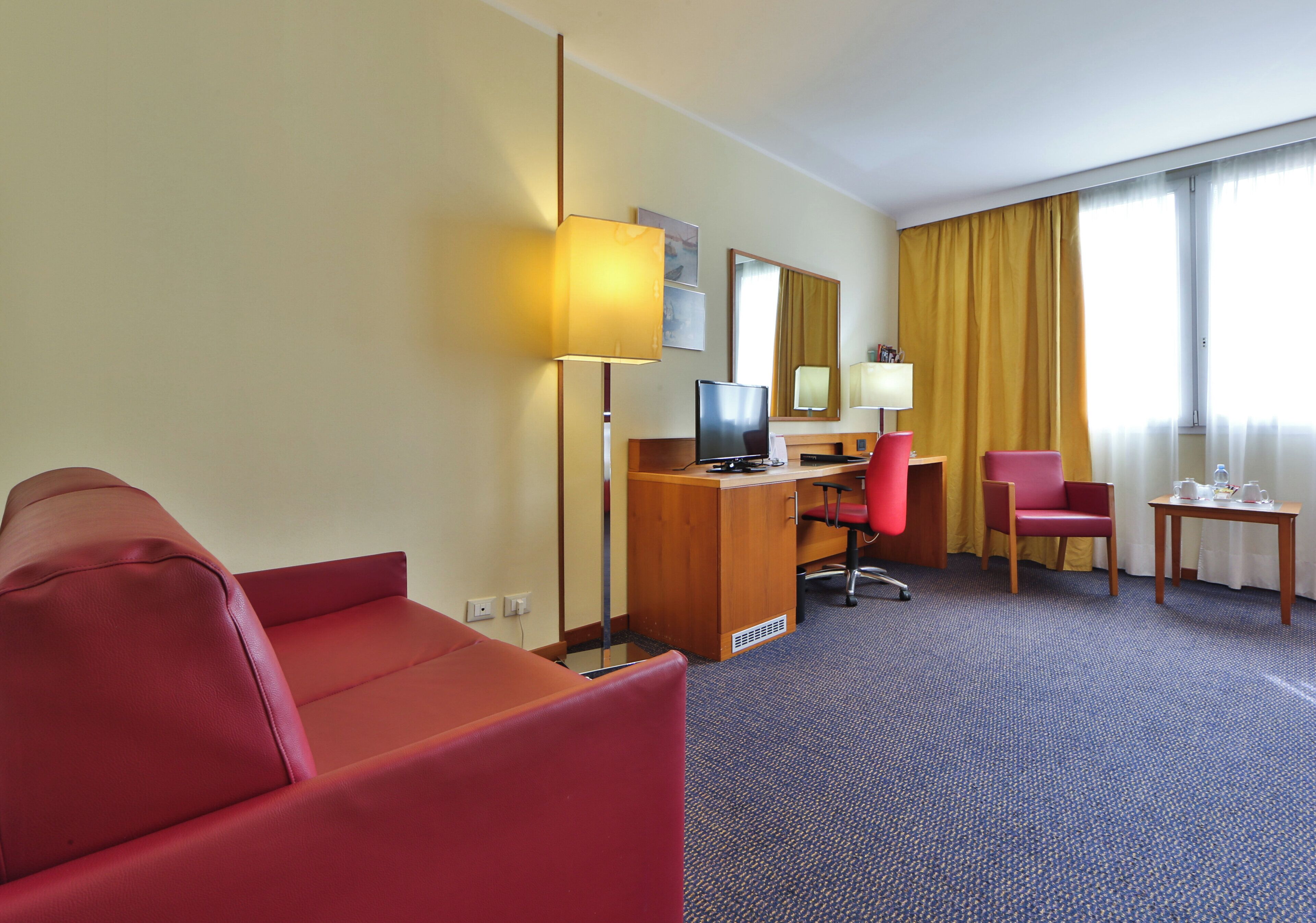 A hotel room with a red sofa, a desk with a computer, a chair, a small table with a lamp, and a large window with curtains.