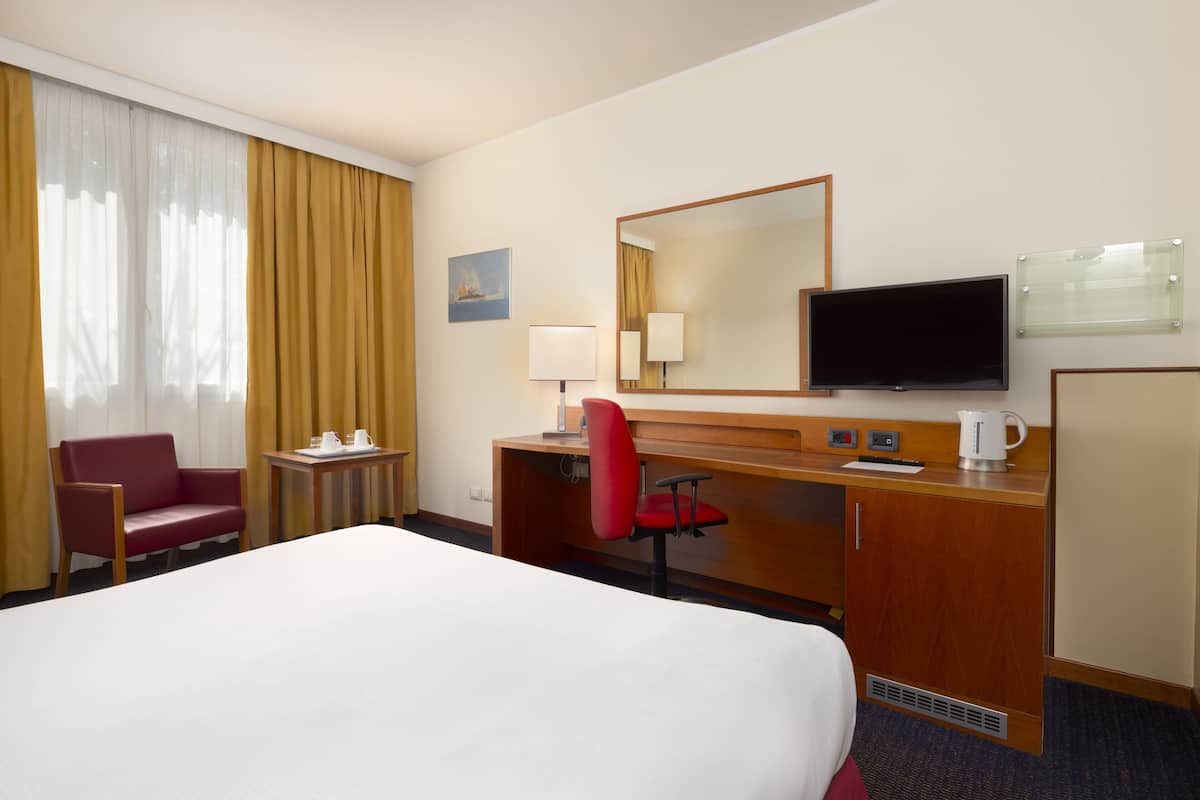 Ein Hotelzimmer mit einem großen Bett, einem Schreibtisch mit Stuhl, einem Fernseher und einem Fenster mit Vorhängen.