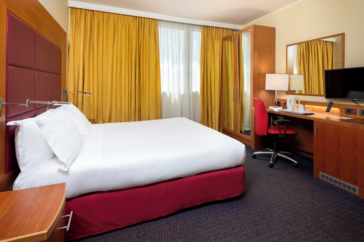 Une chambre d’hôtel avec un grand lit, un bureau avec une chaise, une télévision et une fenêtre avec des rideaux.