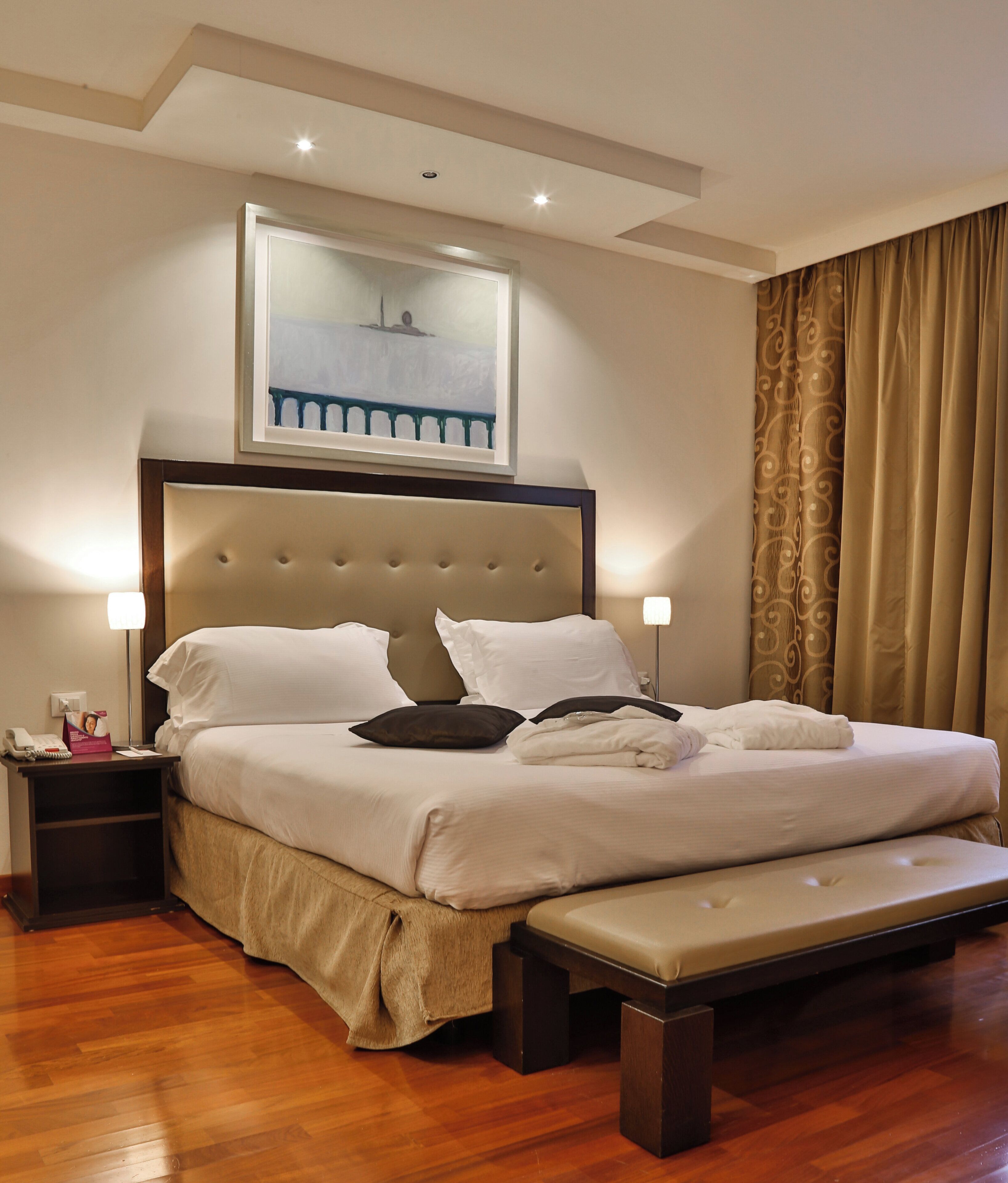 A hotel room with a large bed, bedside lamps, a wooden floor, and a painting above the bed.