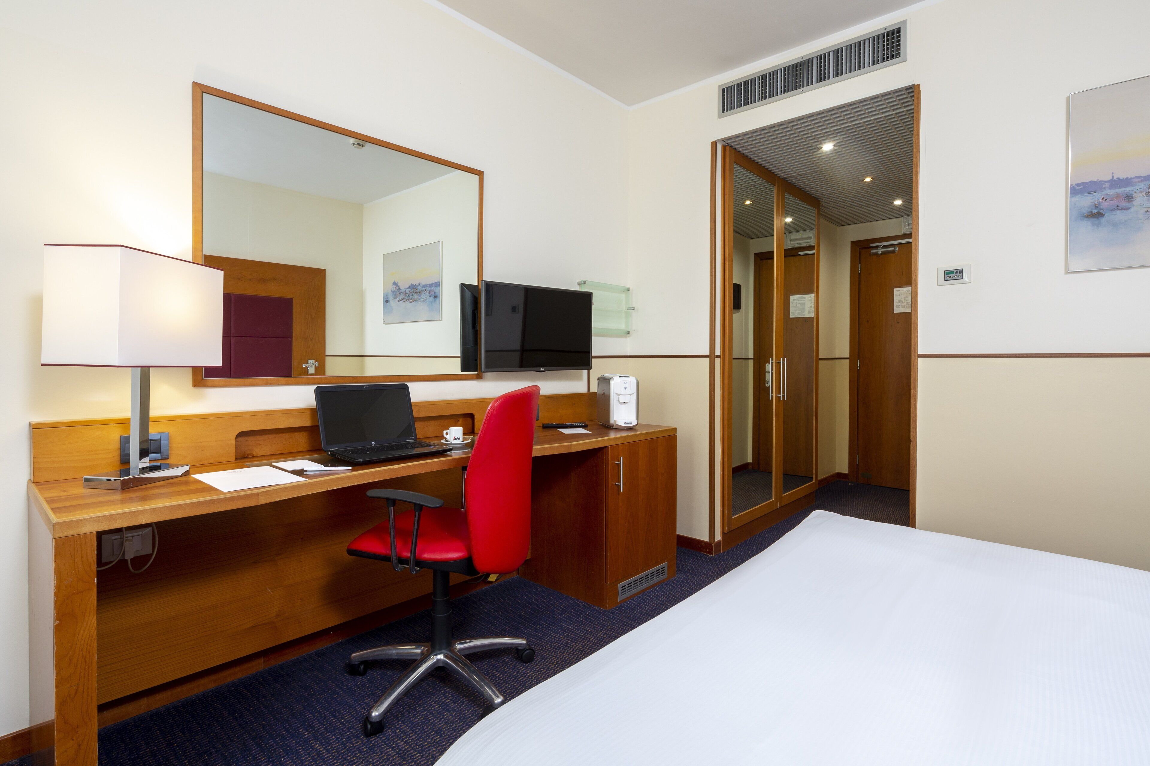 A hotel room with a bed, a desk with a computer, a red chair, a TV, and a large mirror.