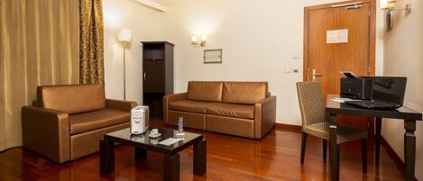 A hotel room with a brown leather sofa, a glass coffee table, a laptop on a desk, and a wooden floor.