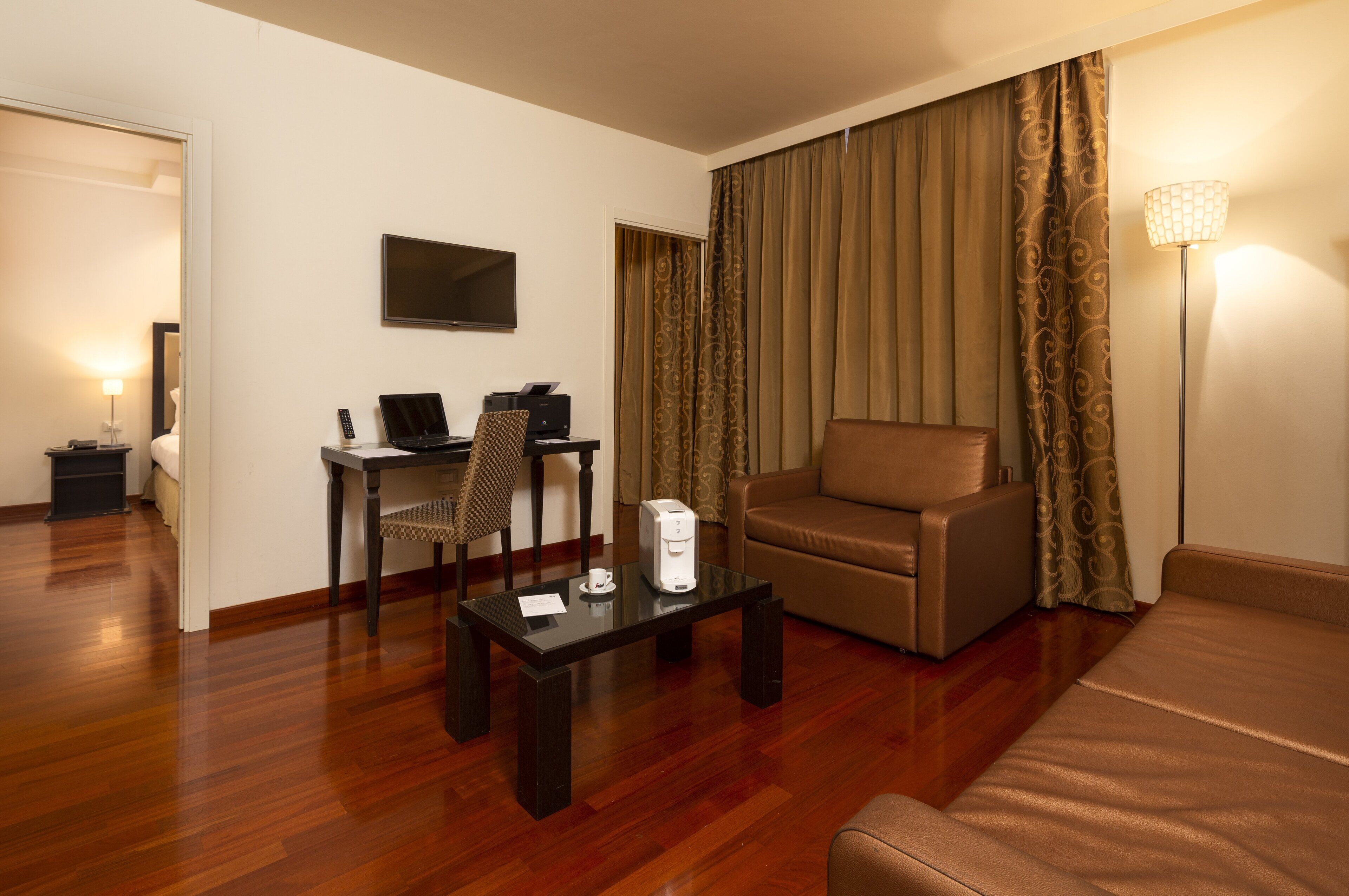 A hotel room with a wooden floor, a sofa, a desk with a chair, a flat-screen TV, and a lamp.