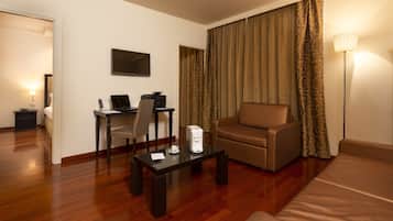 A hotel room with a wooden floor, a sofa, a desk with a chair, a flat-screen TV, and a lamp.