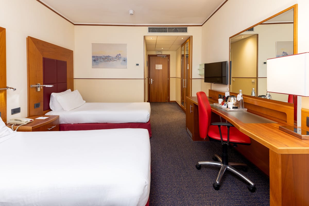 Ein Hotelzimmer mit zwei Betten, einem Schreibtisch mit Stuhl, einem Fernseher, einem Spiegel und einem Bild an der Wand.