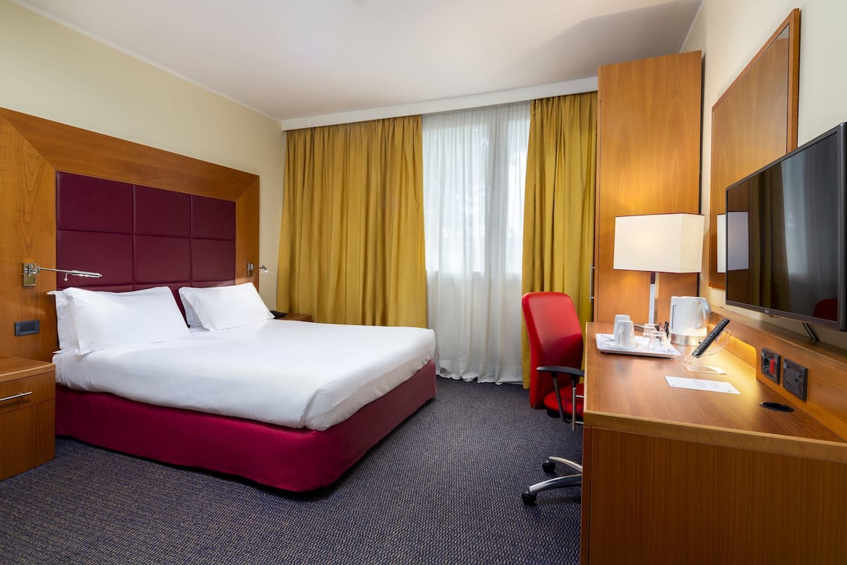 Ein Hotelzimmer mit einem großen Bett, einem Schreibtisch mit Lampe, einem Sessel und einem Fernseher.