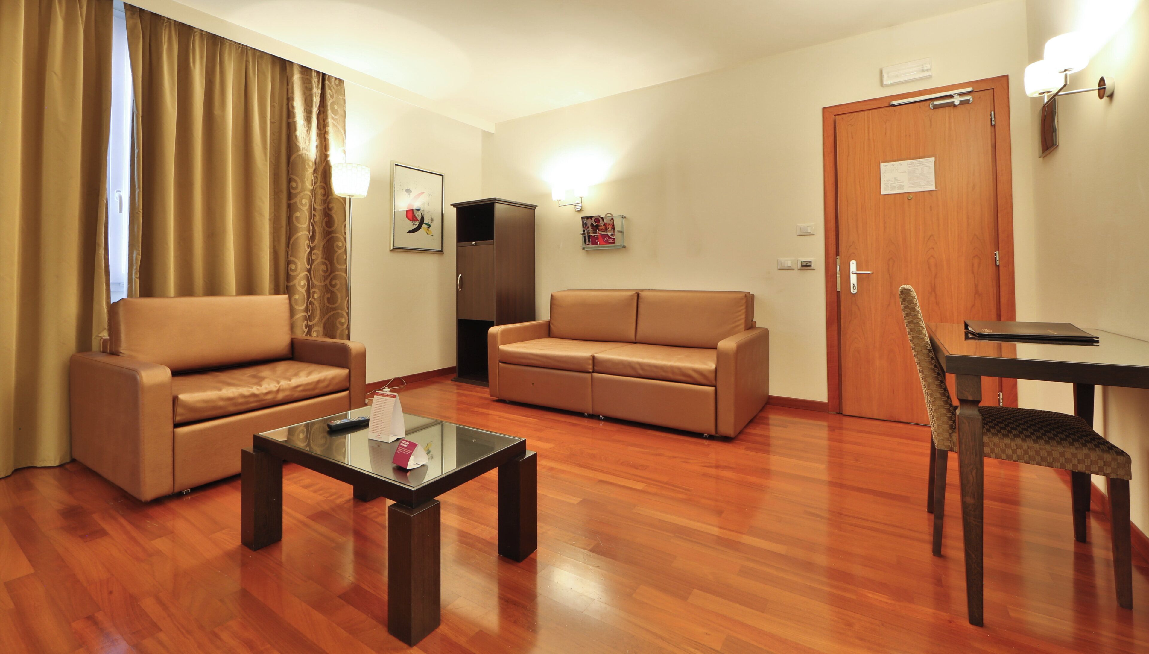 A hotel room with a sofa, a smaller sofa, a glass coffee table, a wooden chair, a desk, and a door.