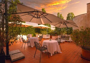 Une terrasse extérieure avec des tables et des chaises rondes, protégée par de grands parasols, entourée de plantes en pot et d’un mur de briques.