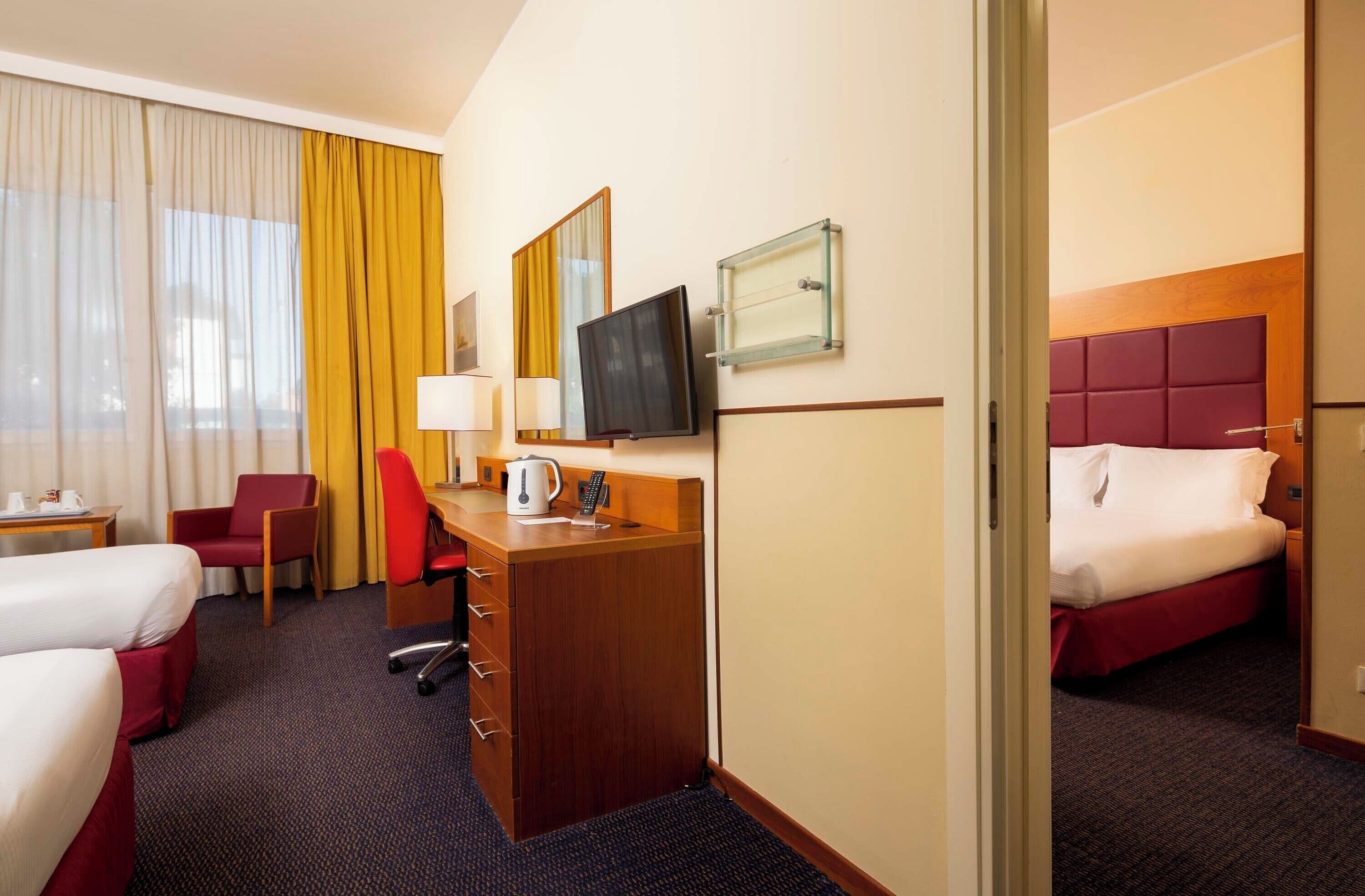 A hotel room with two beds, a desk with a chair, a television, and a window with curtains.