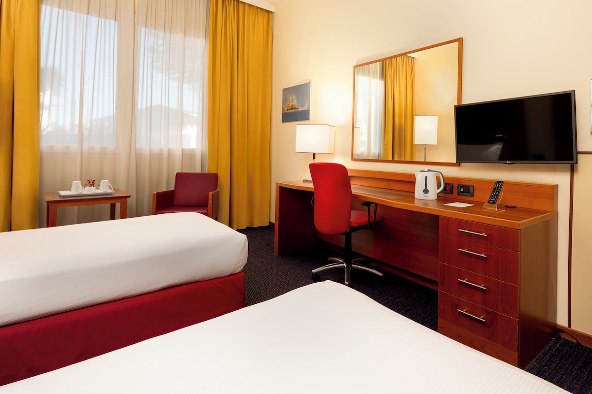 Une chambre d’hôtel avec deux lits, un bureau avec une chaise, une télévision et une fenêtre avec des rideaux.