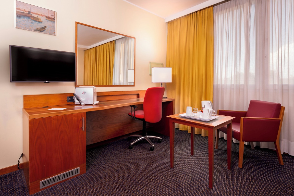 Une chambre d’hôtel avec un bureau en bois, un fauteuil de bureau rouge, une télévision, un miroir, une fenêtre avec des rideaux jaunes et un fauteuil rouge.