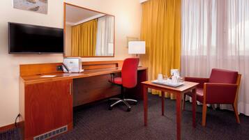 Una habitación de hotel con un escritorio de madera, una silla de oficina roja, un televisor, un espejo, una ventana con cortinas amarillas y suelo alfombrado.