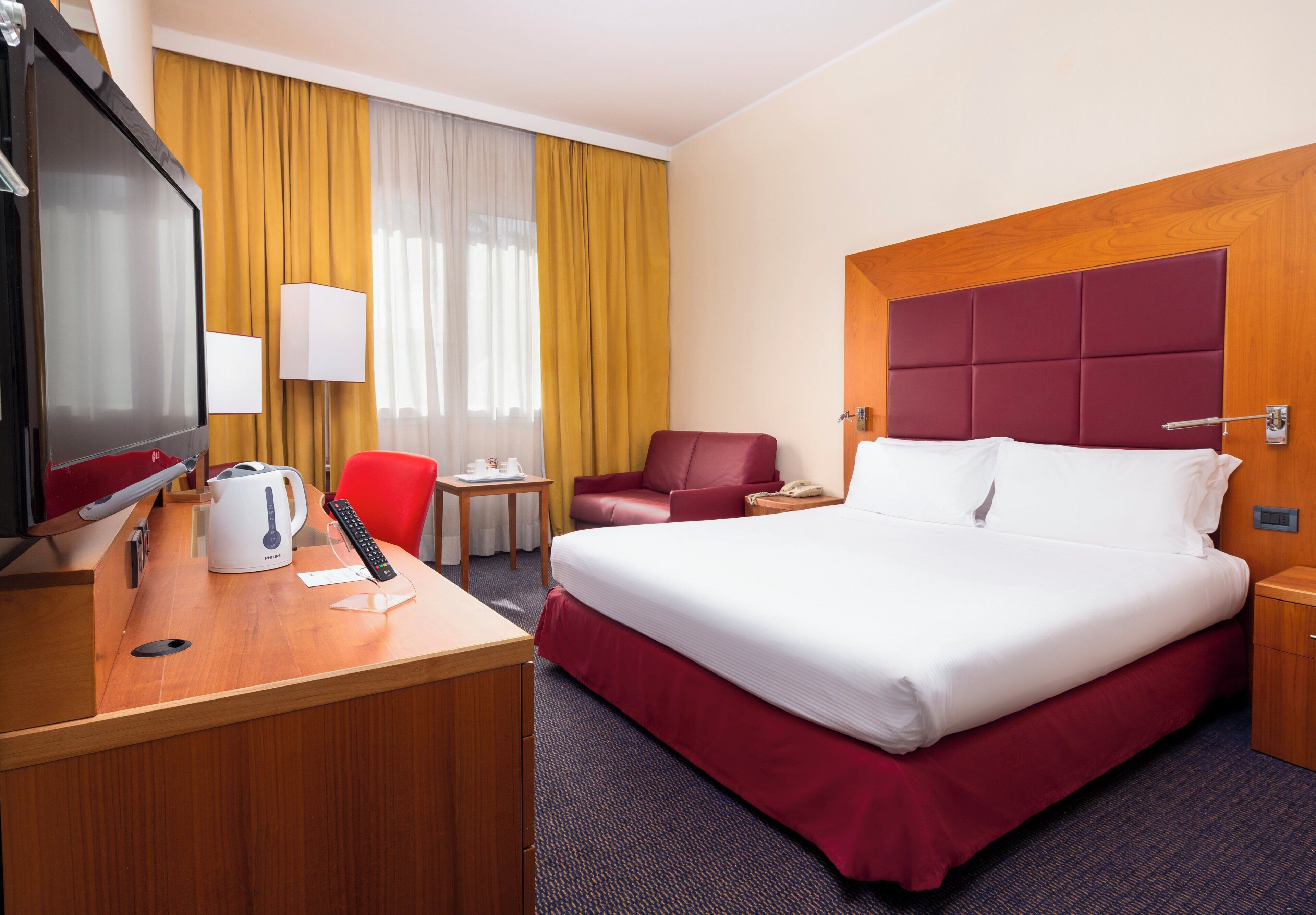 A hotel room with a bed, a desk with a coffee maker, a chair, a TV, and a window with curtains.
