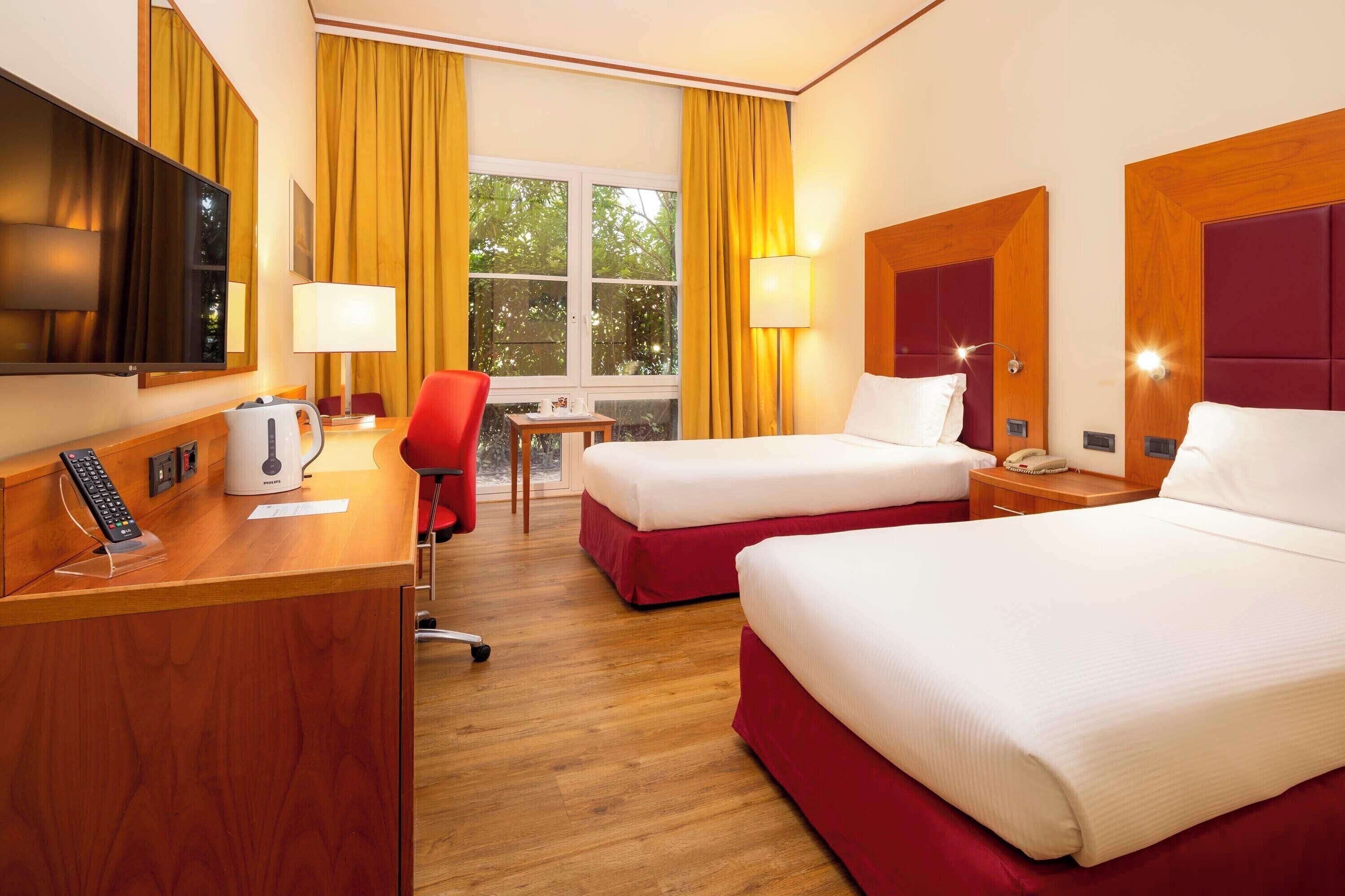 Une chambre d’hôtel avec deux lits, un bureau, une chaise, une télévision et une fenêtre avec des rideaux.