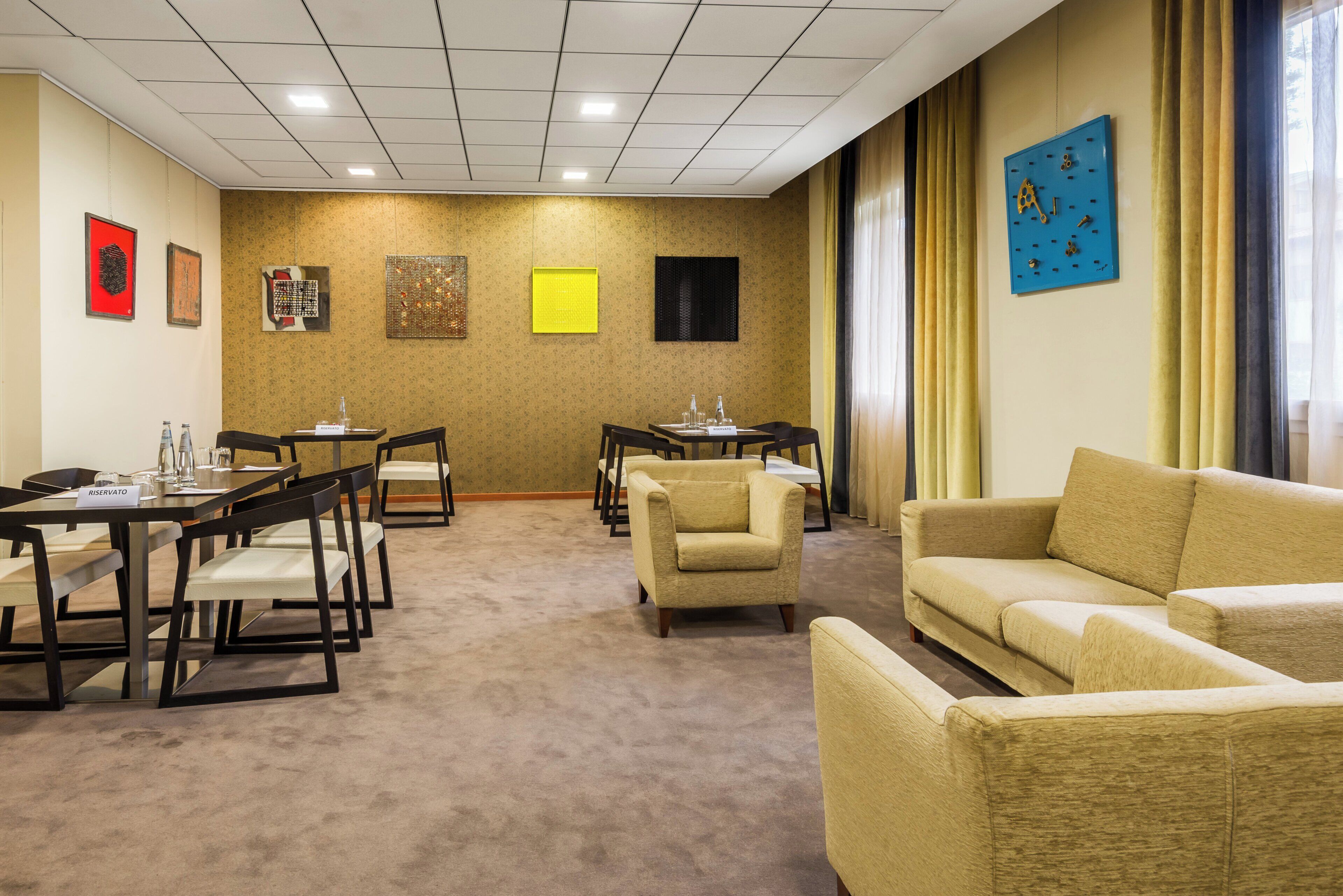 A hotel room with a carpeted floor, a sofa, a dining table with chairs, and artwork on the walls.