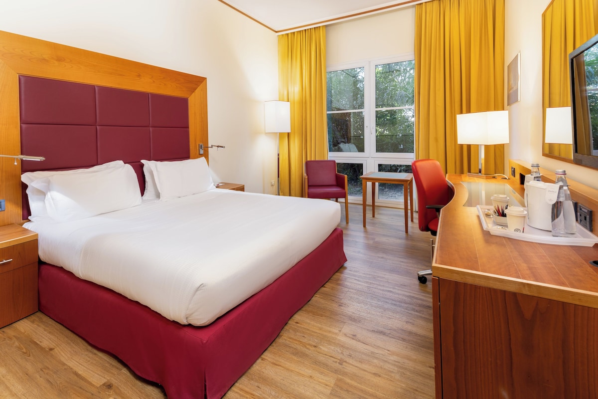 Une chambre d’hôtel avec un grand lit, un bureau avec une chaise, une fenêtre avec des rideaux et une tête de lit en bois.
