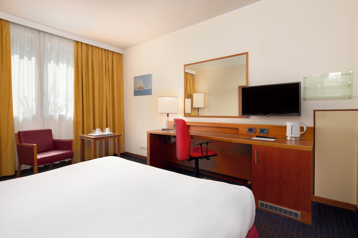 Une chambre d’hôtel avec un grand lit, un bureau avec une chaise, une télévision et une fenêtre avec des rideaux.