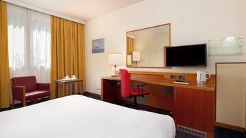 Quarto de hotel com cama, escrivaninha, cadeira, televisão e janela com cortinas.