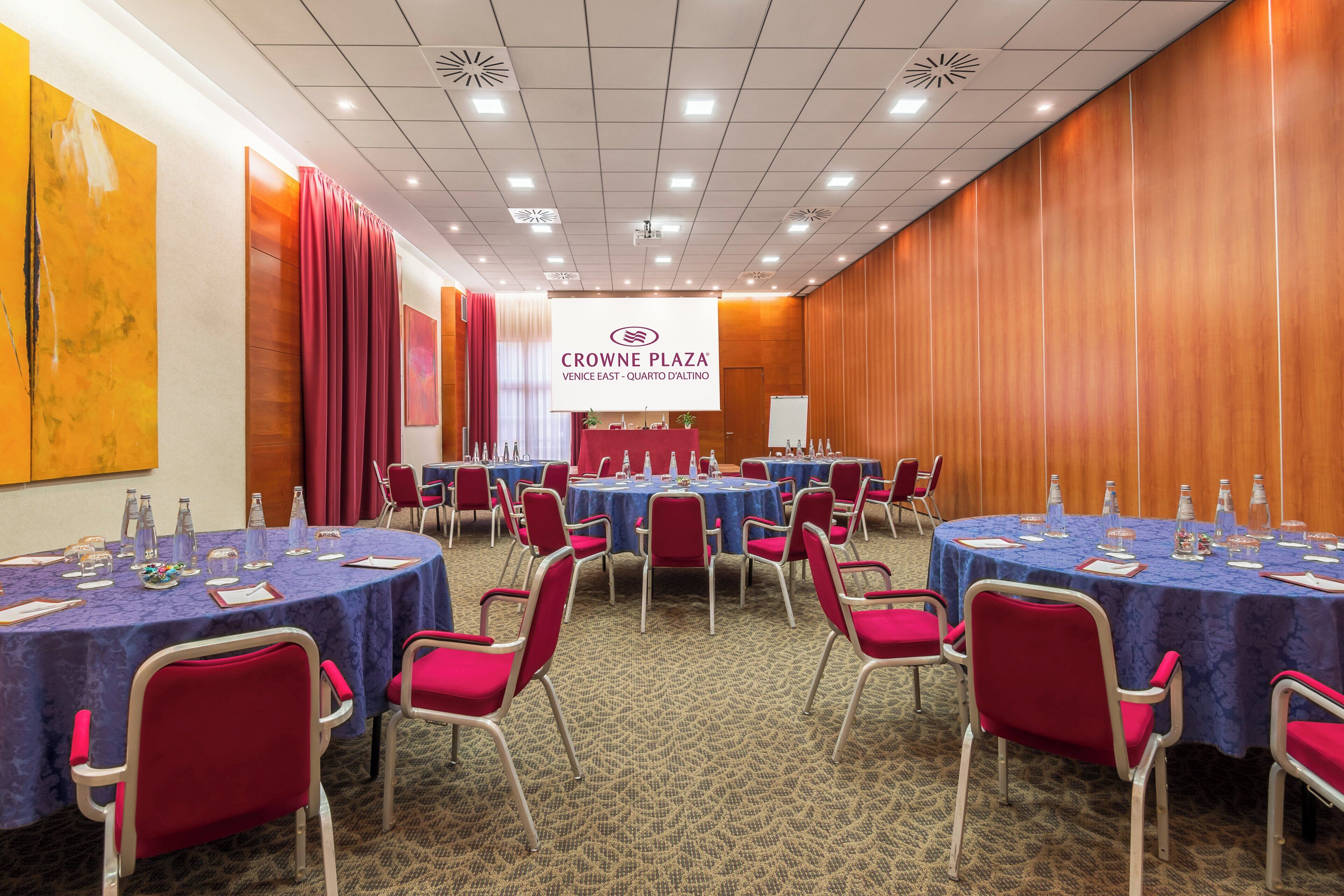 Una sala de conferències a Crowne Plaza, Venice East, amb taules rodones, cadires vermelles i una pantalla de presentació.
