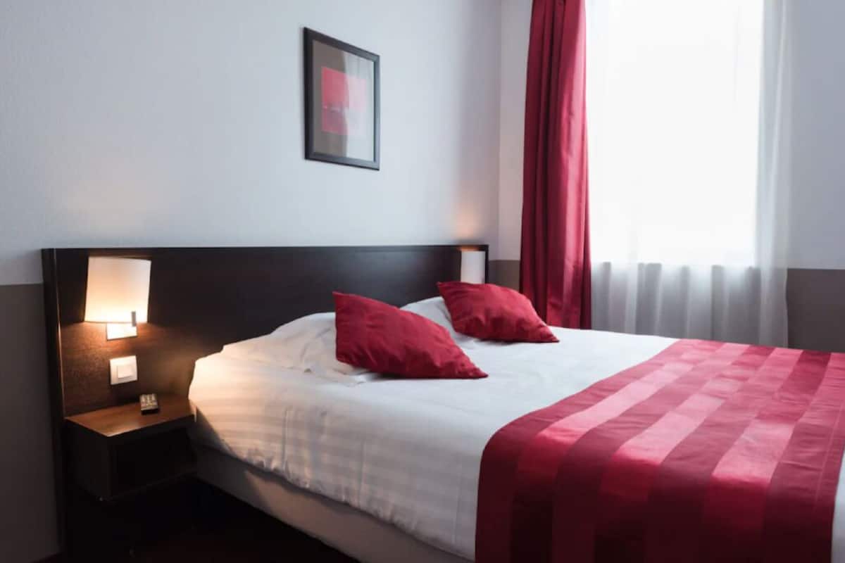 Habitación de hotel con cama, almohadas rojas, mesita de noche con lámpara y un cuadro en la pared.