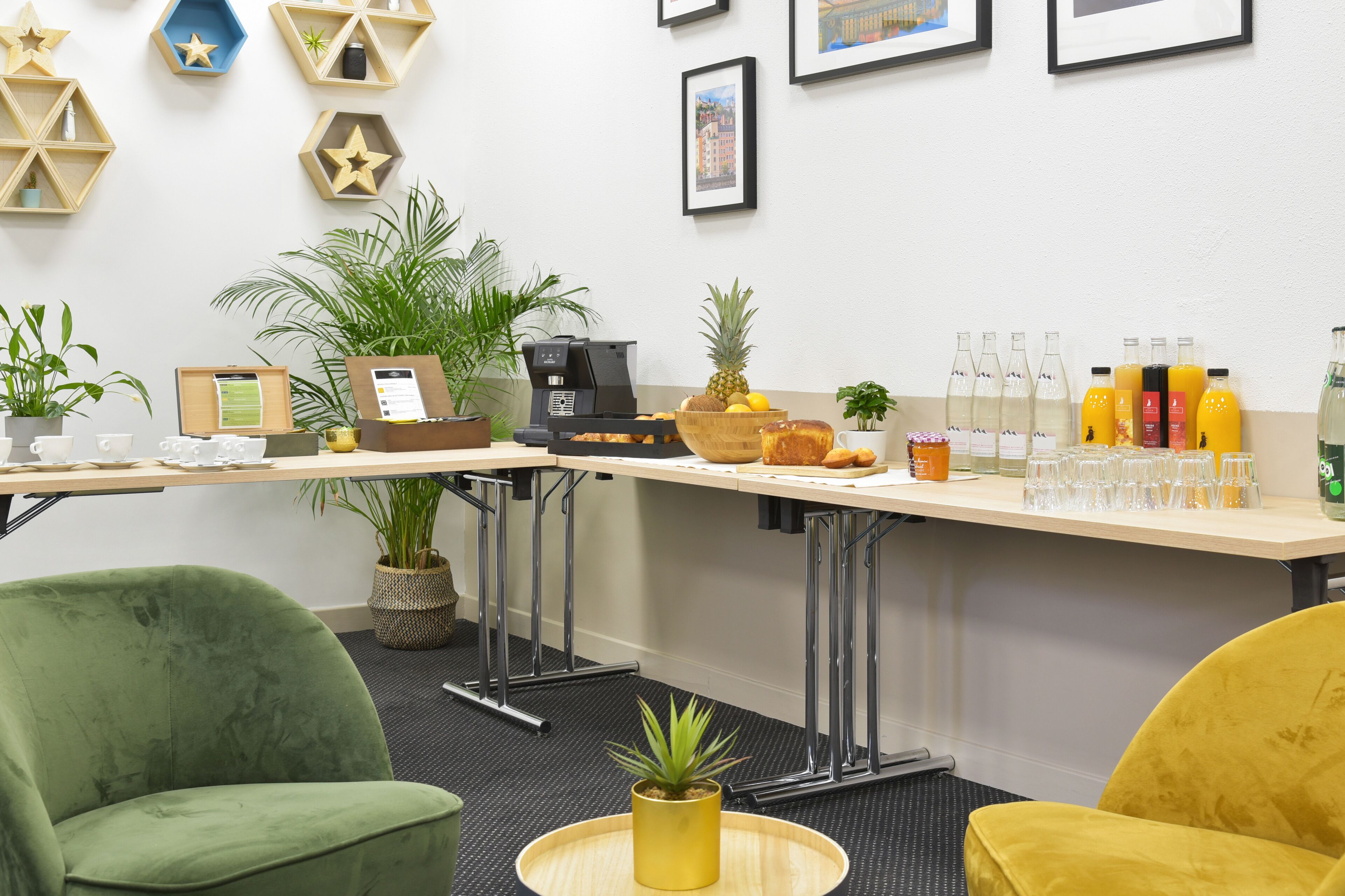 Zona bar moderna con macchina del caffè, frutta fresca e diverse bevande. Sono presenti piante in vaso e quadri appesi al muro.