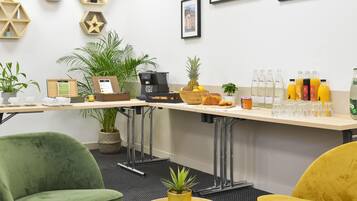 Zona bar moderna con macchina del caffè, frutta fresca e diverse bevande. Sono presenti piante in vaso e quadri appesi al muro.