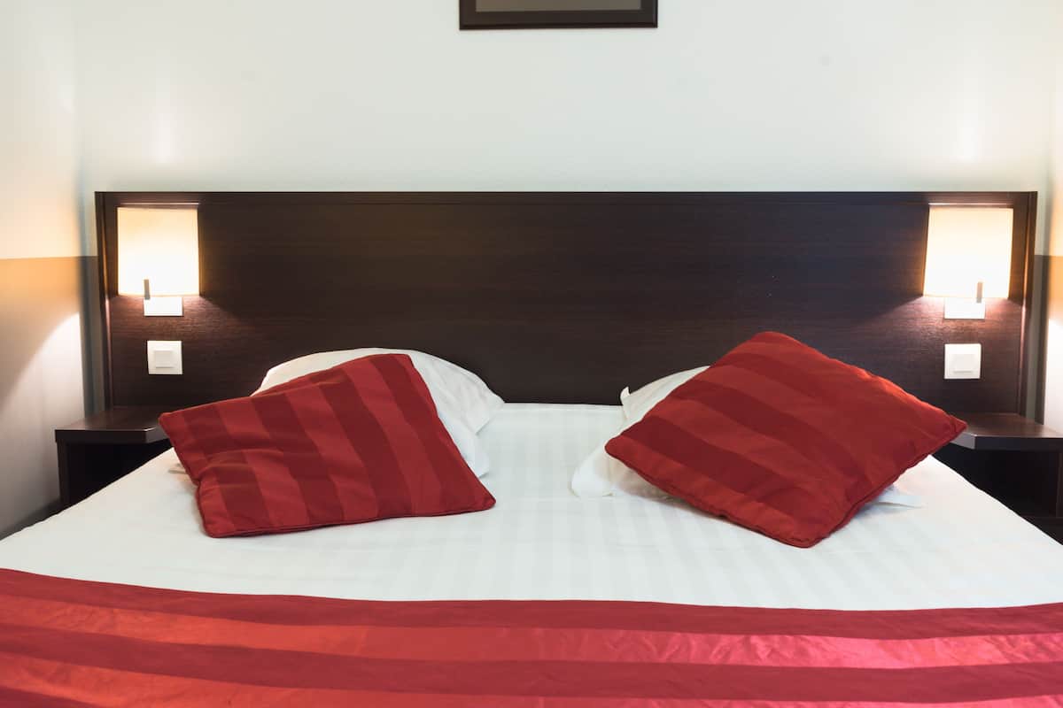 Una cama bien hecha con sábanas blancas y dos almohadas rojas, con dos mesitas de noche y lámparas a cada lado.