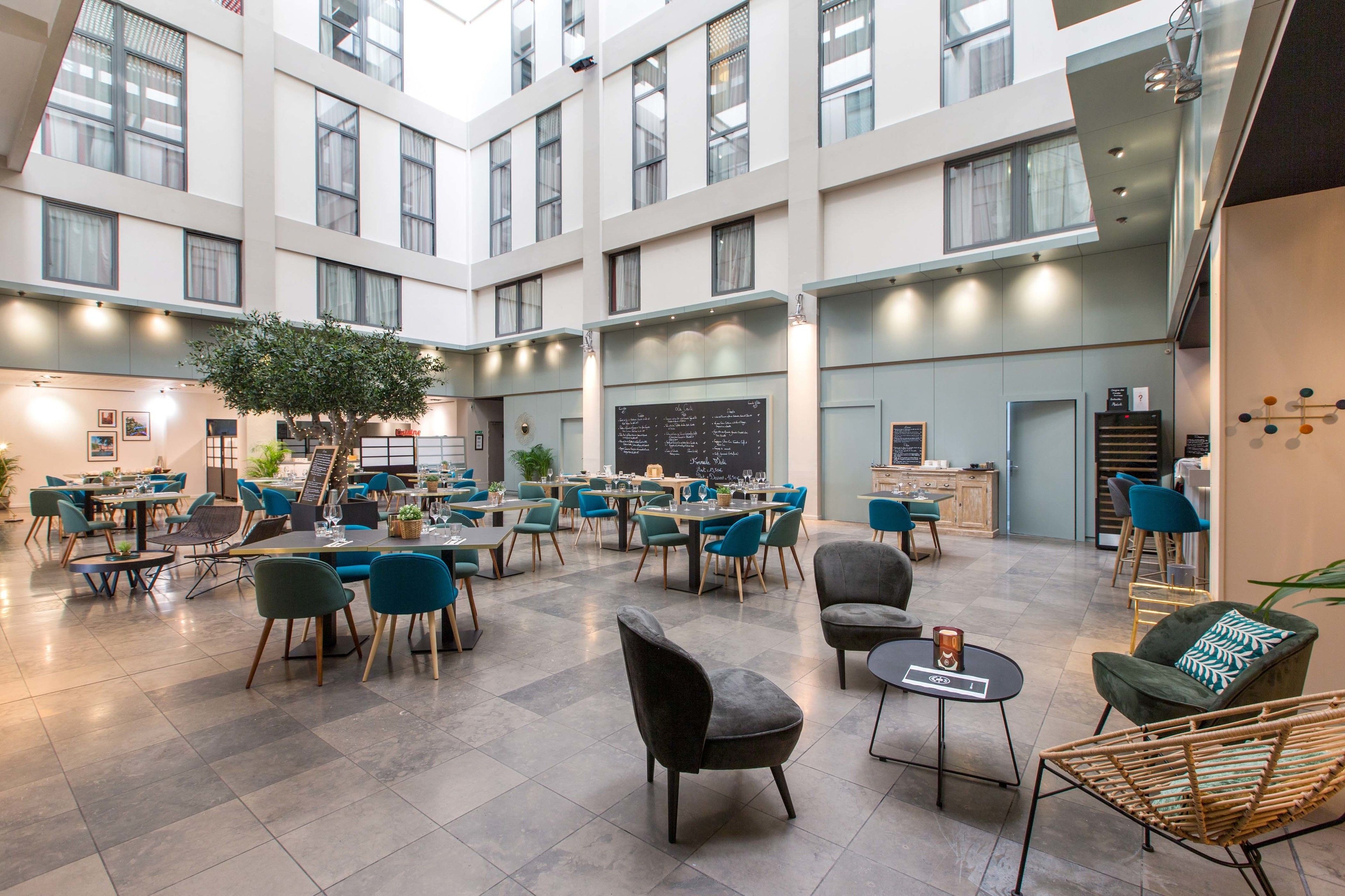 A spacious indoor area with multiple dining tables and chairs, a central seating area with sofas, and a large tree in the middle.