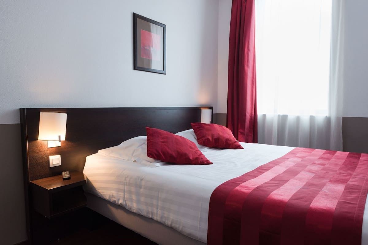 Habitación de hotel con una cama, almohadas rojas, una mesita de noche con lámpara y un cuadro enmarcado en la pared.