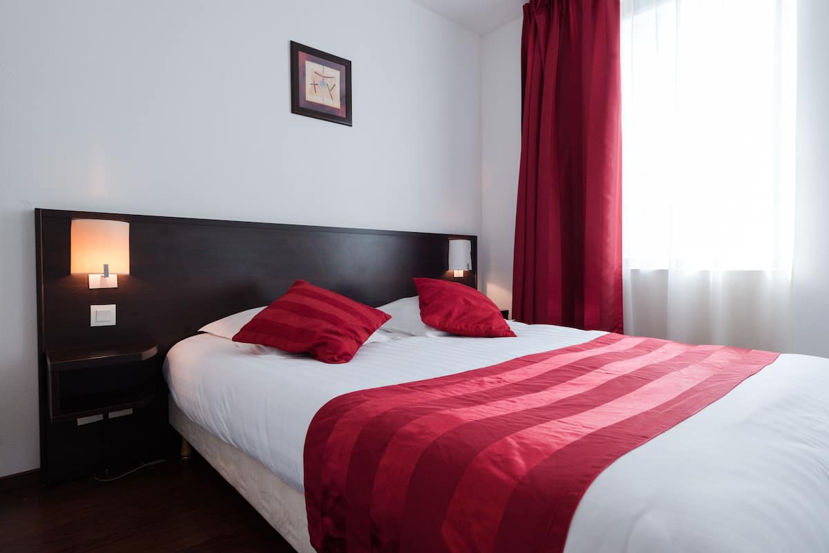 Una cama bien hecha con una colcha blanca y roja a rayas, dos almohadas rojas y un cabecero oscuro. Hay una mesita de noche con una lámpara, y un cuadro enmarcado en la pared sobre la cama.